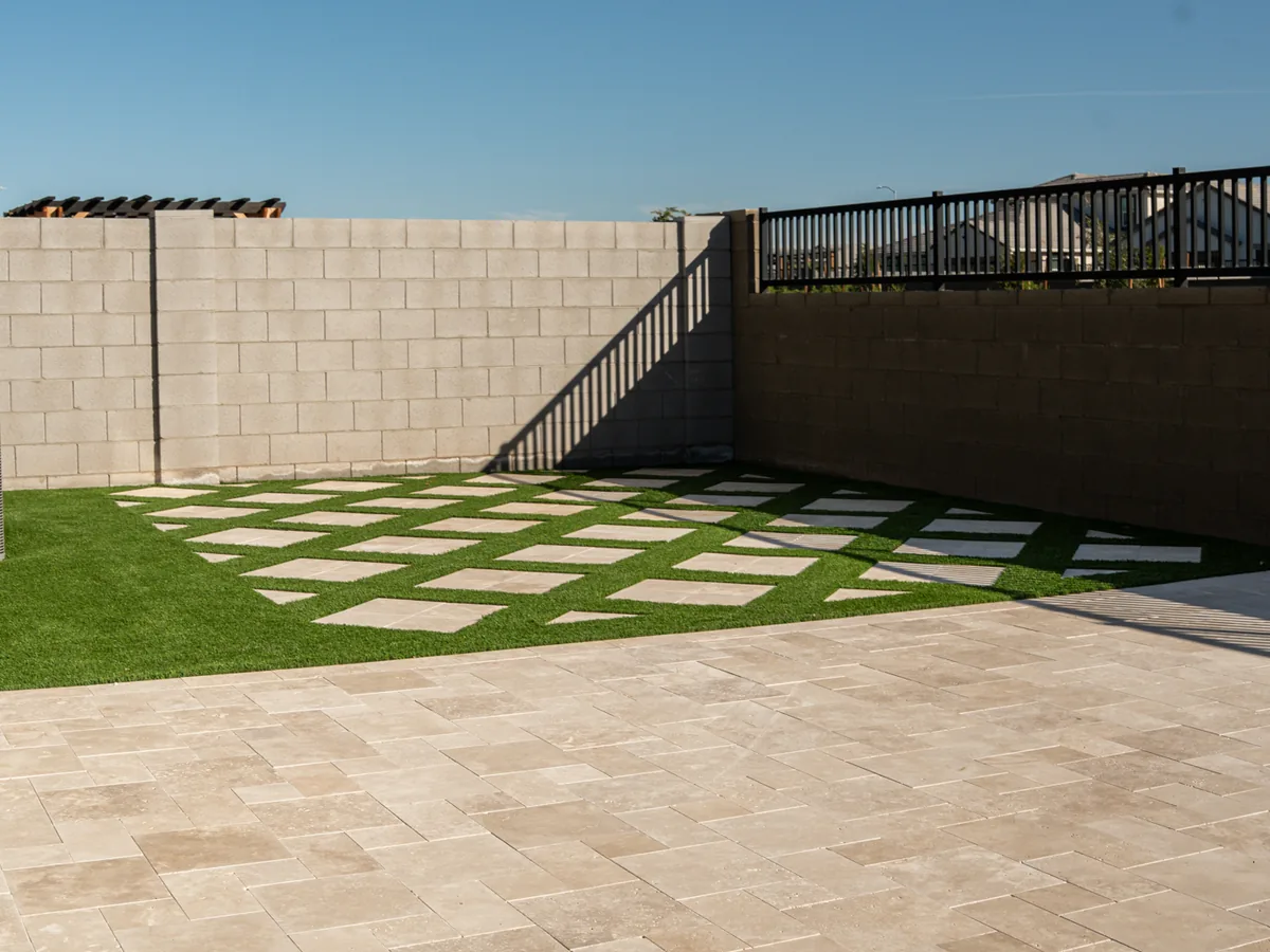 A back yard corner where large square pavers alternate with turf against a block wall. An open turf area runs beside them.