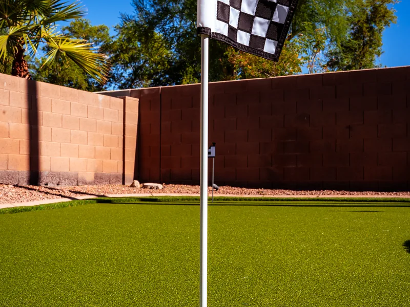 A close view of a flagstick and cup set into a finished putting green. A block wall and trees stand behind it.