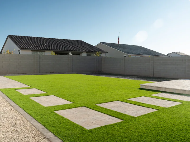 A finished turf back yard. Large paver pads sit flush in the grass. A gravel border runs along the block wall. Single story houses stand behind it.