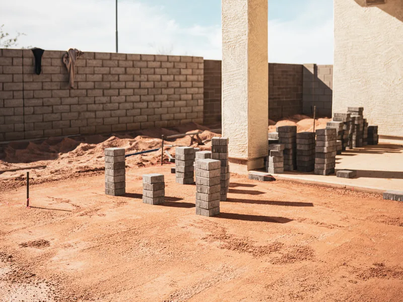 A bare dirt back yard behind a new house. A block wall runs around it. Stacks of pavers sit on the ground. A covered patio is off to one side.