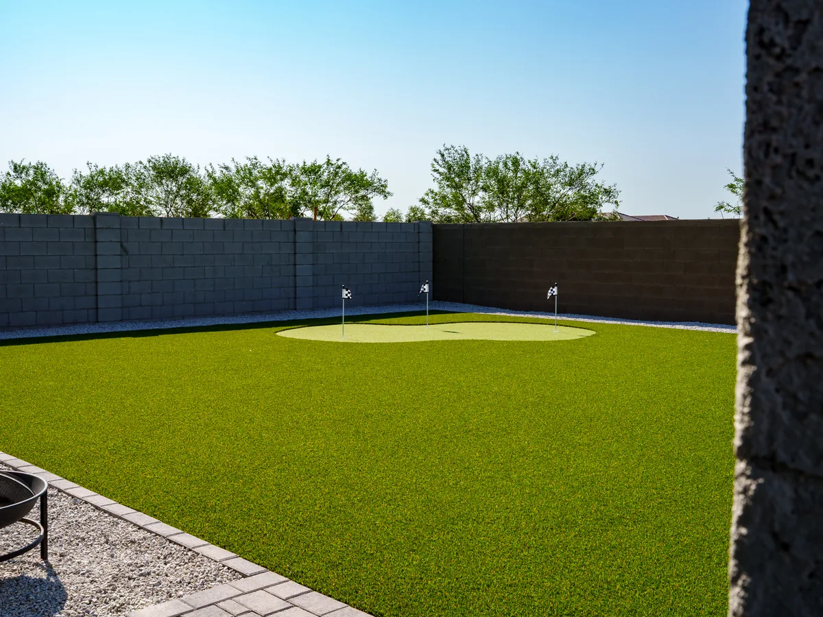 A putting green with flagsticks, set into a large turf back yard. A gravel border runs along the block wall and a paver patio edge sits in front.